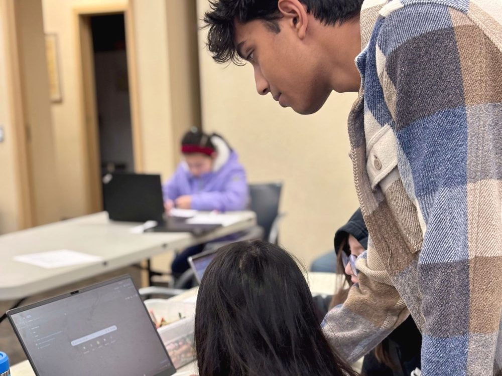 A volunteer leaning in to help with a laptop during a free AI class.