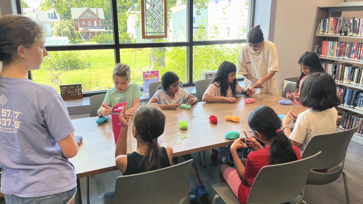 A free crochet class around a long library table beside a wall of windows, coloured yarn in use.